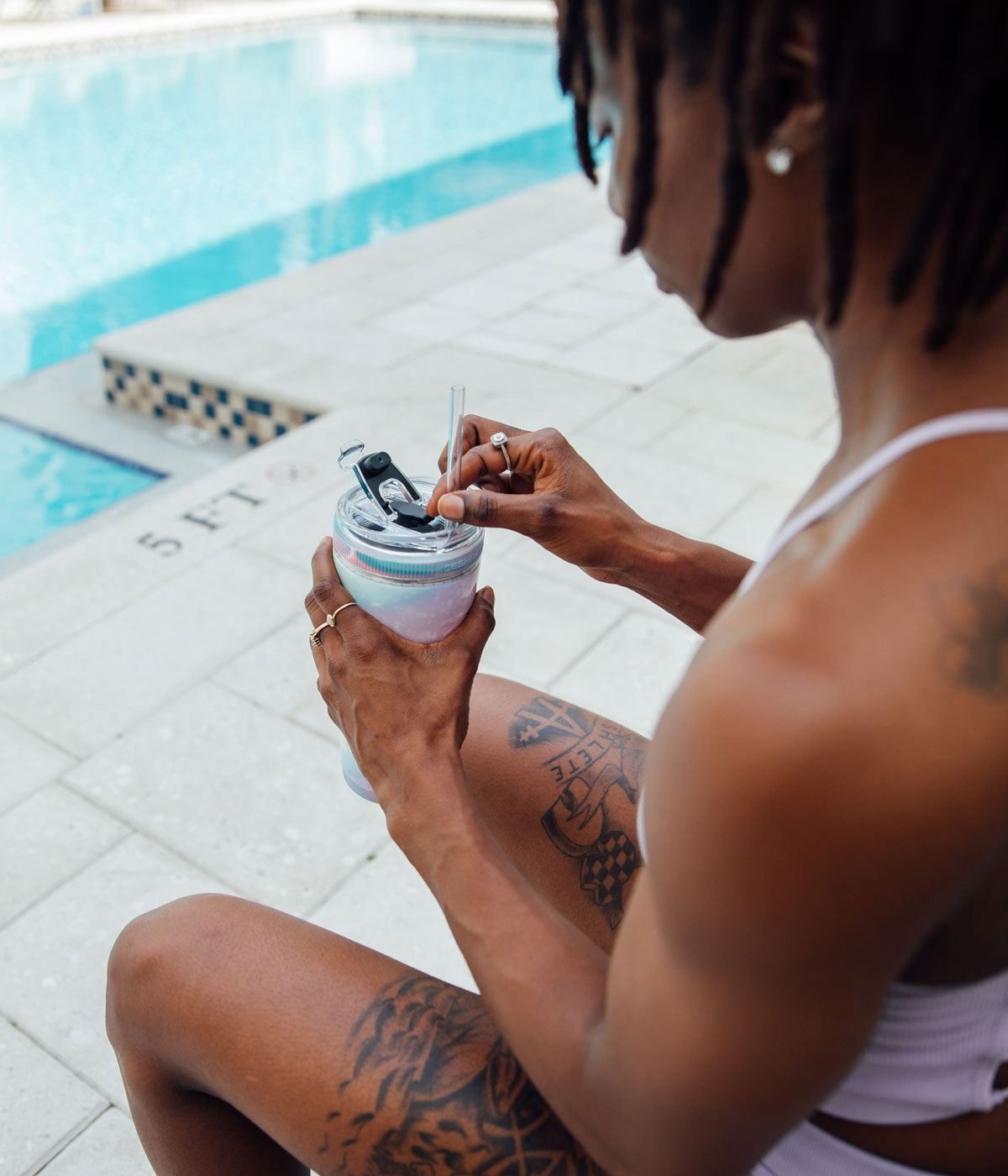 An image of a woman sitting on a bench by a pool. She is holding the Unicorn-colored 26oz Flex Bottle and is actively inserted the straw into the straw hole in the Flex Lid of the Bottle