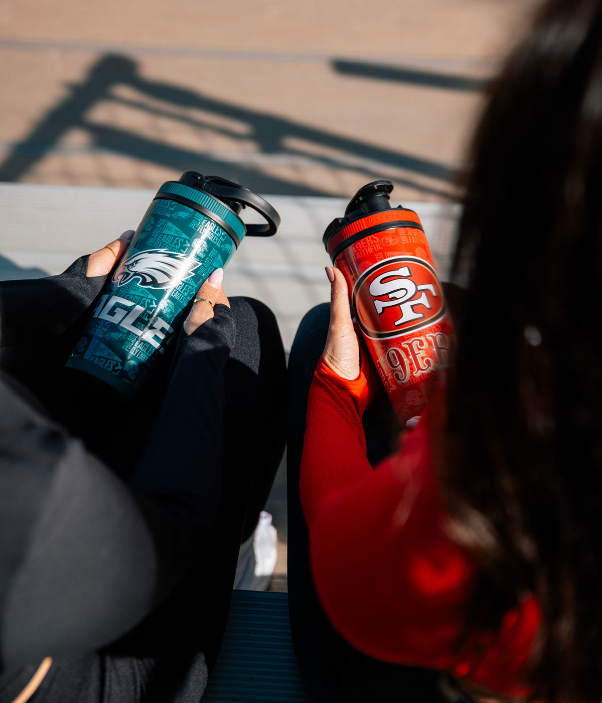 Two girls sitting on bleachers. One girl is holding a Philadelphia Eagles 4D Ice Shaker. The other girl is holding a San Francisco 49ers 4D Ice Shaker.