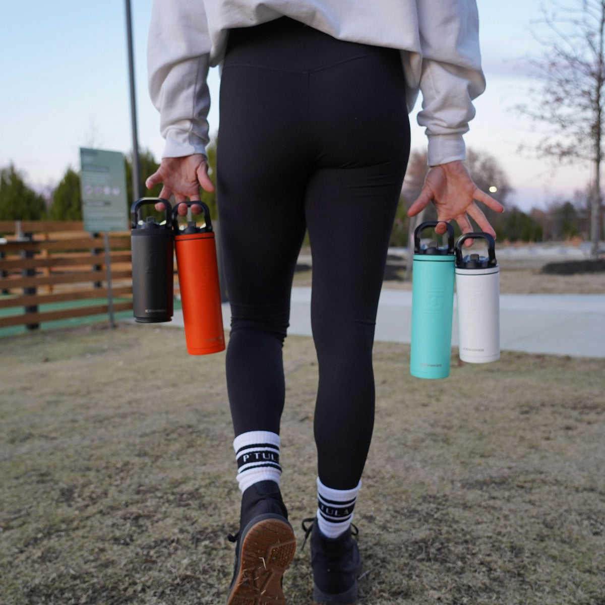 This image shows four different colored Sport Bottles being carried by their handles. A woman is holding them with both of her hands and she is walking away from the camera.